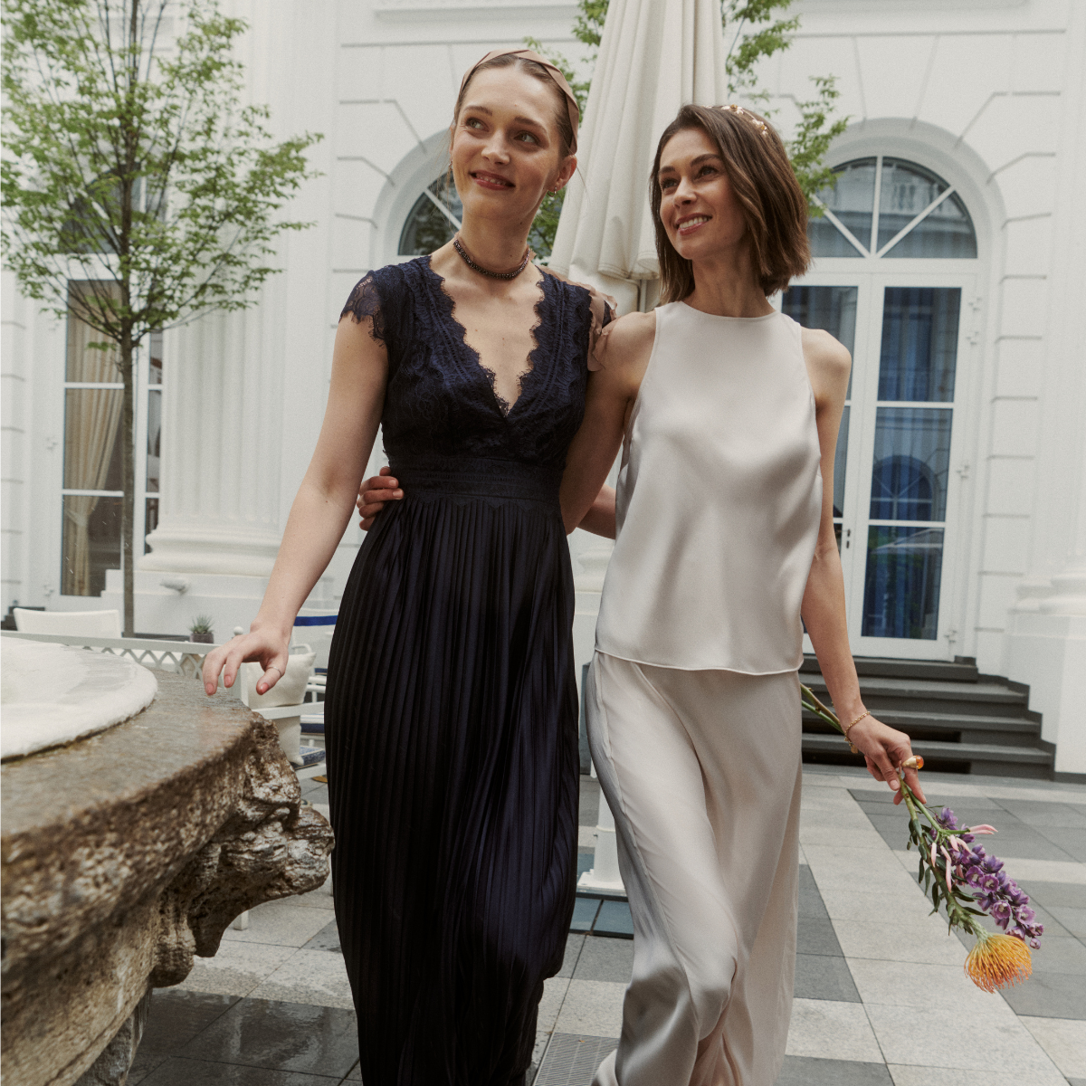 Zwei Damenmodels vor einem hellen Gebäude mit Fenstern. Das linke Model hält in der linken Hand einen kleinen Blumenstrauß mit dem rechten Arm umarmt sie das linke Model. Sie tragen Damen Kleid 'Emelie' in navy und Damen Top 'Laureen' mit Damen Rock 'Fabiola' in champagner aus der Guido Maria Kretschmer Kollektion.