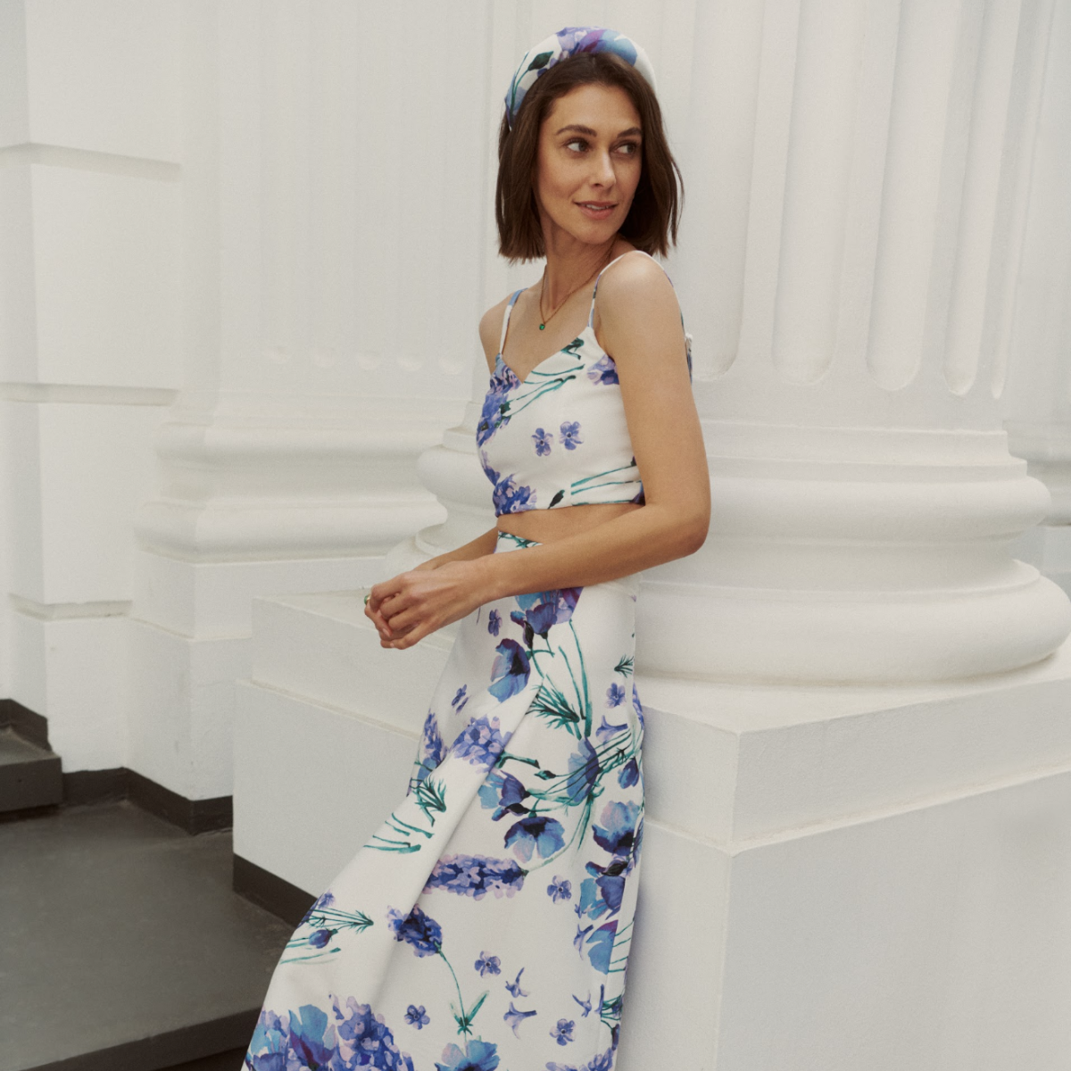 Ein Damenmodel, welches auf einer Treppenstufe steht und sich an einer weißen Säule eines älteren Gebäudes anlehnt. Sie trägt Damen Top 'Ashley' mit Damen Rock 'Caitlin' in weiß mit fliederfarbenen Blumenprint aus der Guido Maria Kretschmer Kollektion.