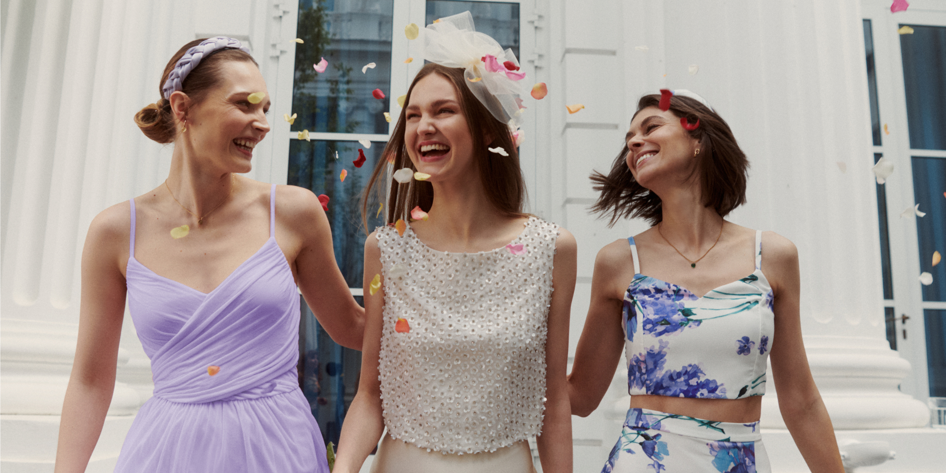 Drei Damenmodels vor einem hellen Gebäude mit Fenstern, in der Luft schwebt buntes Konfetti. Die Damen tragen Damen Kleid 'Cathleen' in lavendel, Damen Rock 'Fabiola' in champagner, Damen Top 'Ashley' und Damen Rock 'Caitlin' in weiß mit lavendelfarbenen Blumenprint aus der Guido Maria Kretschmer Kollektion.