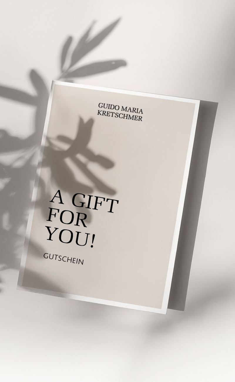 Eine Gutscheinkarte mit der Aufschrift: Guido Maria Kretschmer "A GIFT FOR YOU!" Gutschein. Die Karte liegt auf einem hellen Hintergrund und es ist der Schatten einer Pflanze zu erkennen, welcher sich über die Gutscheinkarte wirft.