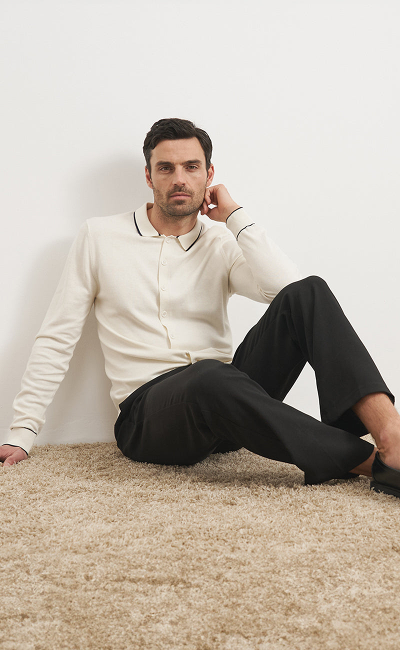 Ein Herrenmodel vor einfarbigem hellen Hintergrund. Das Model Sitzt auf einem Hochfloor Teppich in beige. Er trägt Herren Cardigan 'Torge' in weiß mit navy Details am Kragen und an den Ärmeln. Außerdem trägt er Herren Hose 'Jonte' in navy. Beide Styles stammen aus der Guido Maria Kretschmer Kollektion.