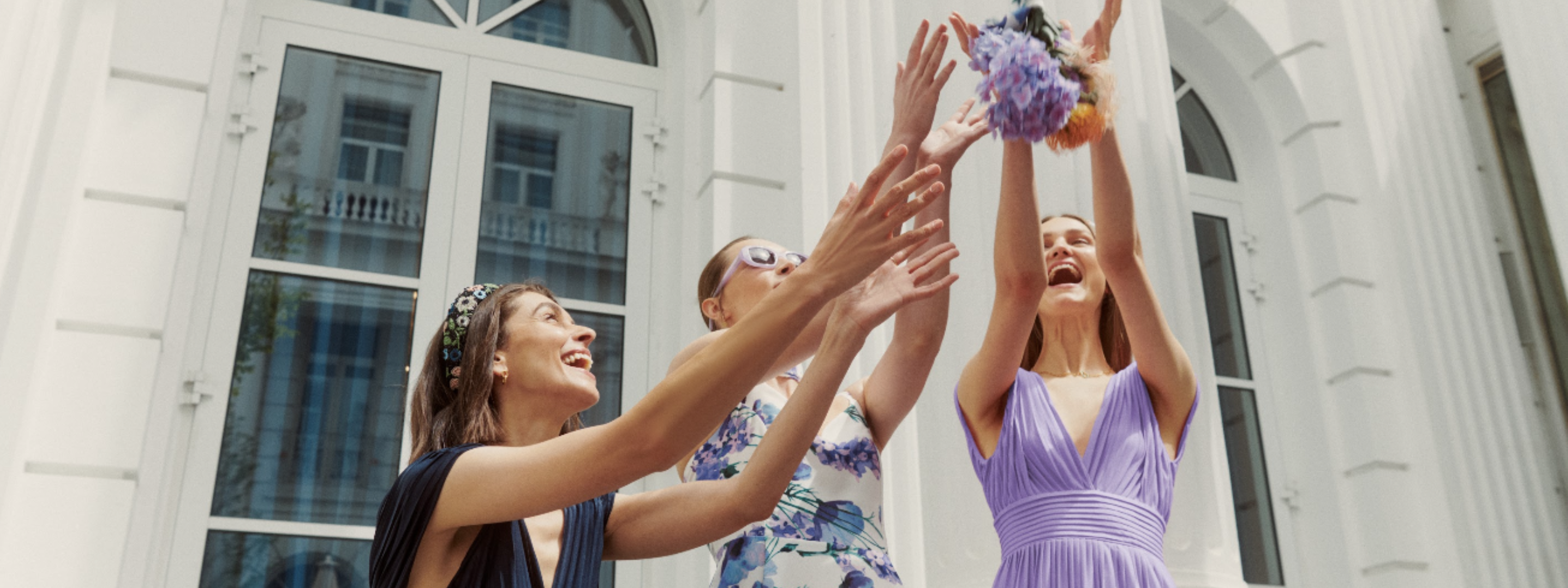 Drei Damenmodels vor einem hellen Gebäude mit Fenstern, welche einen Blumenstrauß fangen. Sie tragen Damen Kleid 'Linnea' in navy, Damen Kleid 'Carola' in weiß mit fliederfarbenen Blumenprint und Damen Kleid 'Linnea' in lavendel aus der Guido Maria Kretschmer Kollektion.