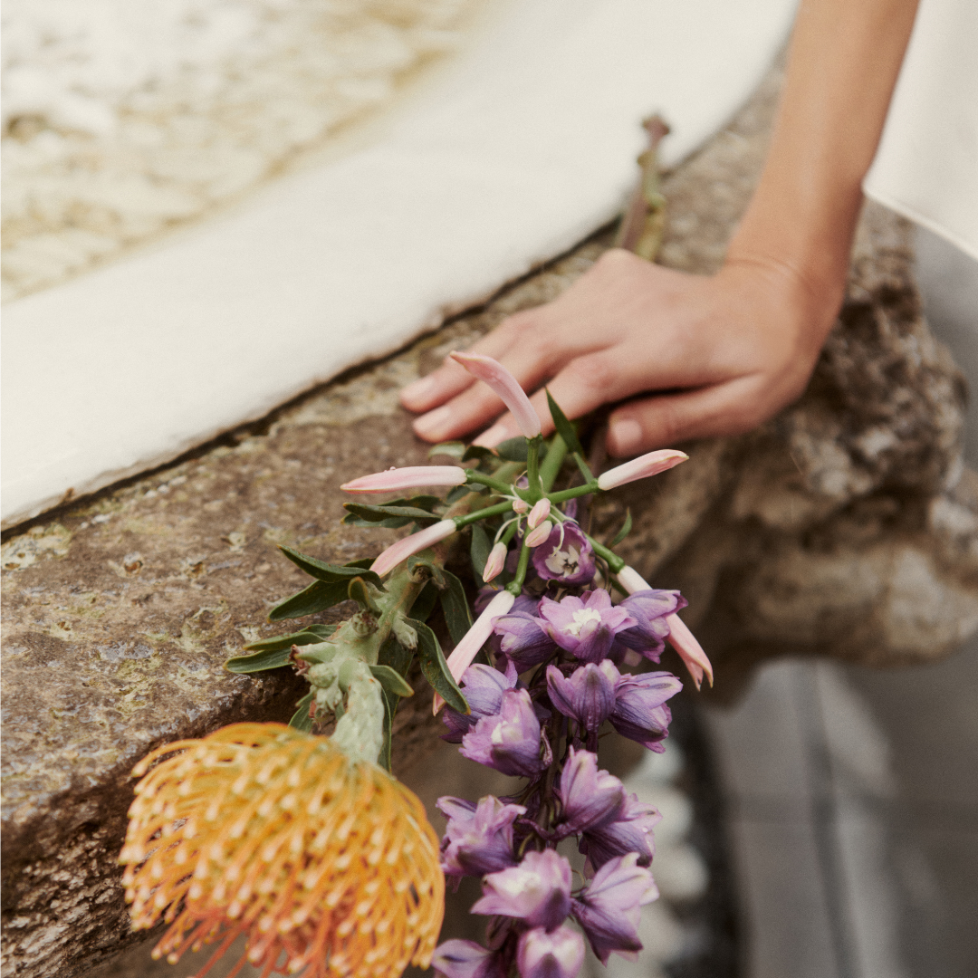 Nahaufnahme verschieden farbiger Blumen, darunter violette Blüten und rosa Knospen. Sie liegen auf einer Steinfläche und werden von einer Hand festgehalten.