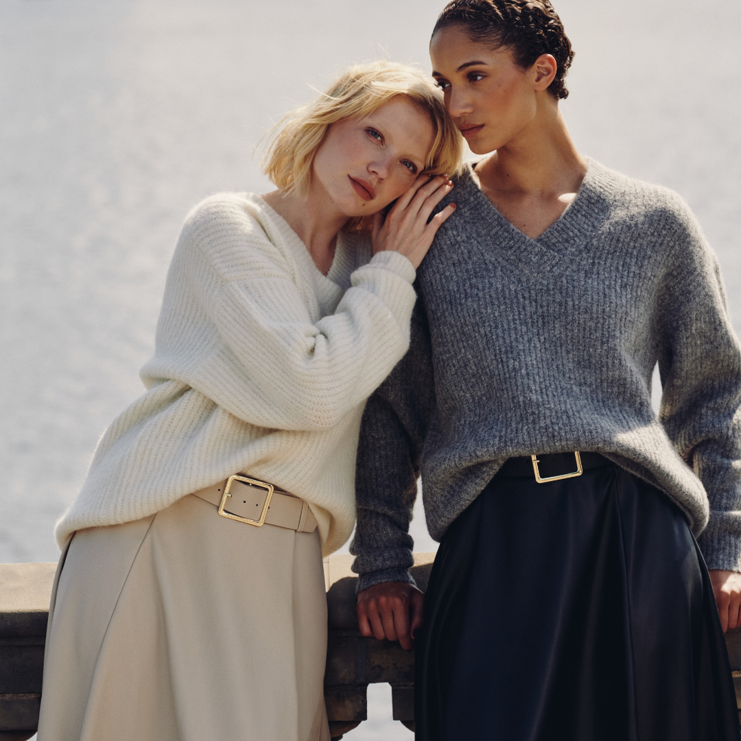 Zwei Damenmodels stehen vor einer Skulptur in der Stadt Hamburg. Das linke Model trägt einen hellbeigen Pullover und einen hellbeigen Rock und das rechte Model einen hellgrauen Pullover und einen dunkelblauen Rock. Das Outfit stammt aus der Guido Maria Kretschmer Kollektion.