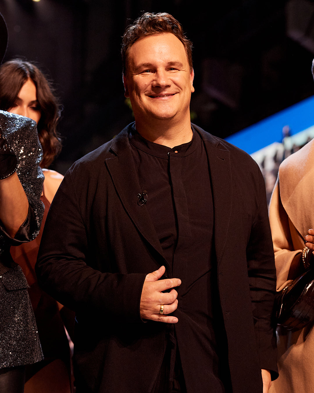 Guido Maria Kretschmer steht zwischen den Models der About You Fashion Week in Berlin 2021. Er trägt einen schwarzen Anzug und lächelt.