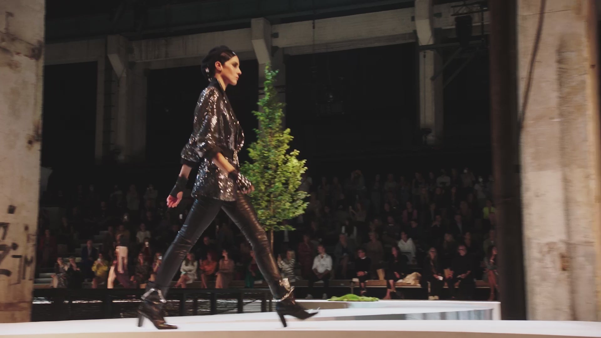 Auf dem Video läuft ein Damenmodel einen Laufsteg vor Publikum entlang. Es handelt sich um die About You Fashion Week in Berlin 2021. Das Model trägt einen Blazer, welcher mit Pailletten in lila besetzt ist, der BH darunter ist leicht zu sehen, dazu trägt sie eine glänzende schwarze Hose.
